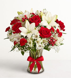 Fields of Europe Christmas Bouquet of red roses and white lilies with a red ribbon in a clear vase on a light gray background