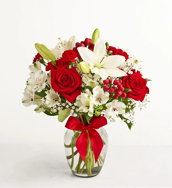 Fields of Europe Christmas Bouquet of red roses and white lilies with a red ribbon in a clear vase on a white background