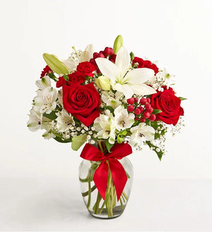 Fields of Europe Christmas Bouquet of red roses and white lilies with a red ribbon in a clear vase on a white background