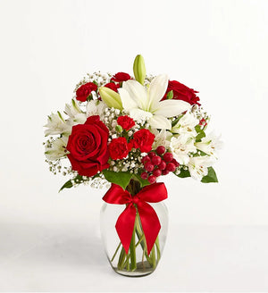 Fields of Europe Christmas Bouquet of red roses and white lilies in a clear vase with a red ribbon on a white background