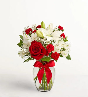 Fields of Europe Christmas Bouquet of red roses and white lilies in a clear vase with a red ribbon on a light gray background