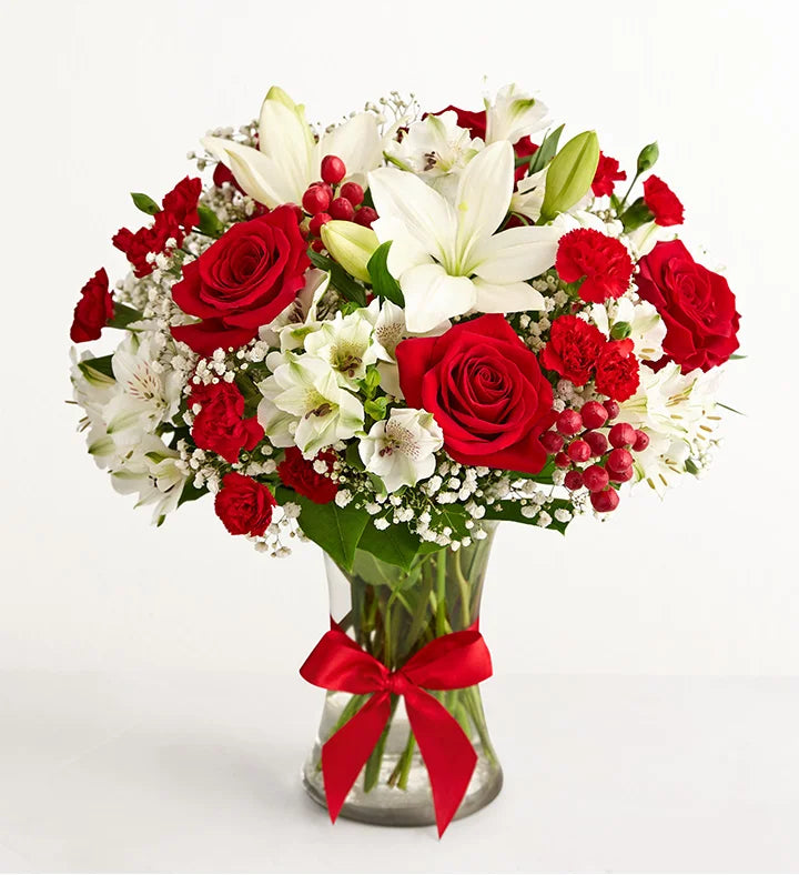 Fields of Europe Christmas Bouquet of red roses, white lilies, and red berries in a clear vase with a red ribbon on a white background.