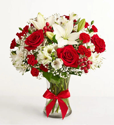 Fields of Europe Christmas Bouquet of red roses, white lilies, and red berries in a clear vase with a red ribbon on a white background. Fields of Europe Christmas Bouquet of red roses, white lilies, and red berries in a clear vase with a red ribbon on a white background.