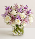 White roses with pink alstroemeria, purple stock, white carnations and baby's breath arranged in a clear glass vase.