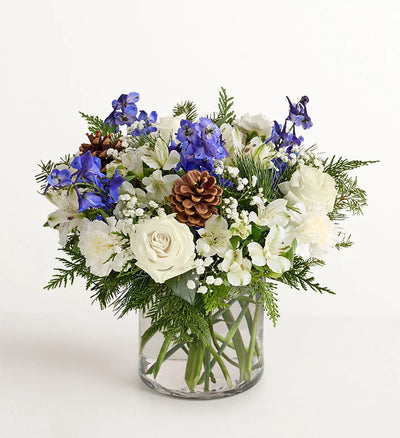 Winter Wishes Bouquet of flowers with white roses, blue delphinium, and pinecones in a clear vase on a white background Winter Wishes Bouquet of flowers with white roses, blue delphinium, and pinecones in a clear vase on a white background