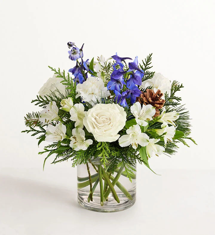Winter Wishes Bouquet of flowers with white roses, blue delphinium, and greenery in a clear vase on a white background