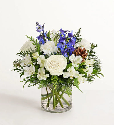 Winter Wishes Bouquet of flowers with white roses, blue delphinium, and greenery in a clear vase on a white background Winter Wishes Bouquet of flowers with white roses, blue delphinium, and greenery in a clear vase on a white background