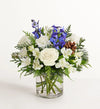 Winter Wishes Bouquet of flowers with white roses, blue delphinium, and greenery in a clear vase on a white background
