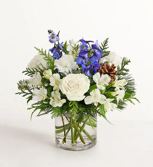 Winter Wishes Bouquet of flowers with white roses, blue delphinium, and greenery in a clear vase on a white background