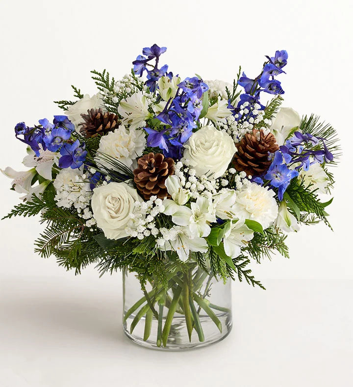 Winter Wishes Bouquet of flowers with blue and white flowers, pine cones, and greenery in a clear vase on a white background