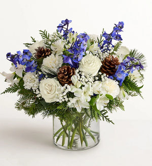 Winter Wishes Bouquet of flowers with blue and white flowers, pine cones, and greenery in a clear vase on a white background