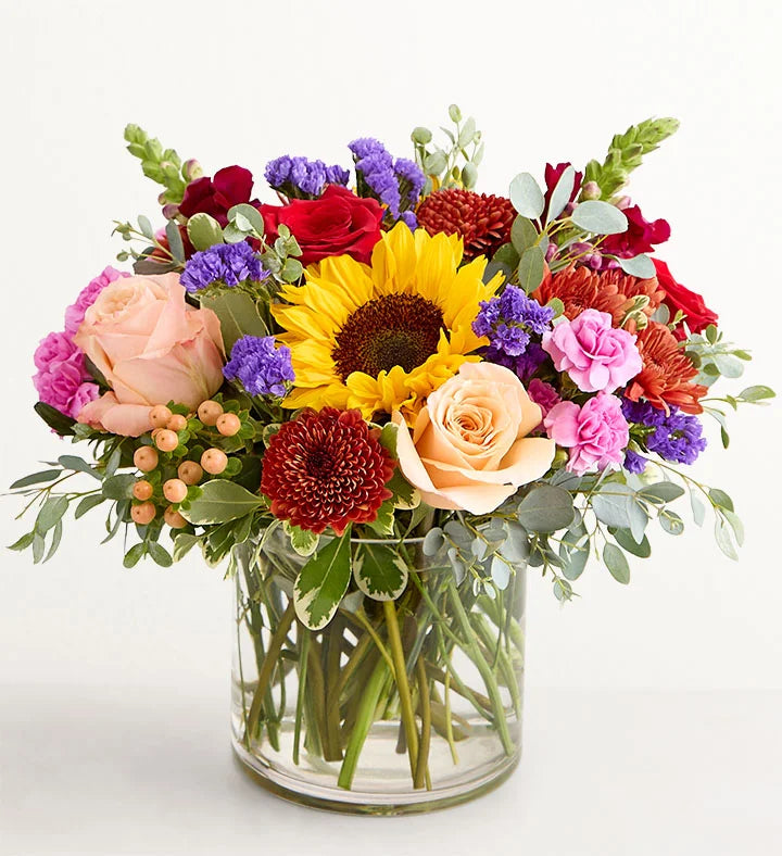 Harvest Melody Bouquet Colorful flower bouquet with sunflowers, roses, and other flowers in a clear vase on a white background