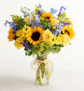 Bouquet of yellow sunflowers and roses with blue accents in a clear vase on a white background