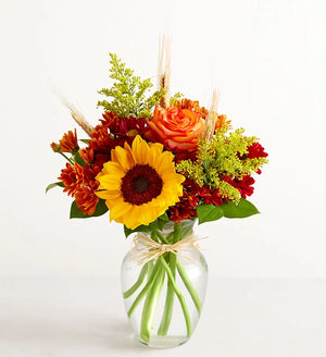 Bouquet of flowers with sunflowers, roses, and greenery in a clear vase on a white background
