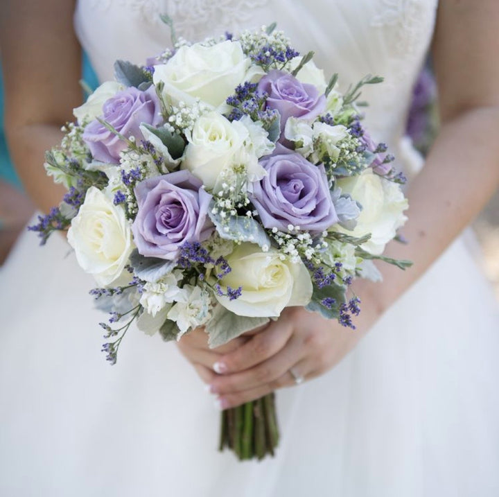 A bride holds a round bridal bouquet composed of soft lavender and white roses, accented with sprigs of baby's breath, purple statice, and dusty miller leaves. The bouquet is neatly arranged and the bride's white dress provides a clean, elegant background for the pastel colors of the flowers.
