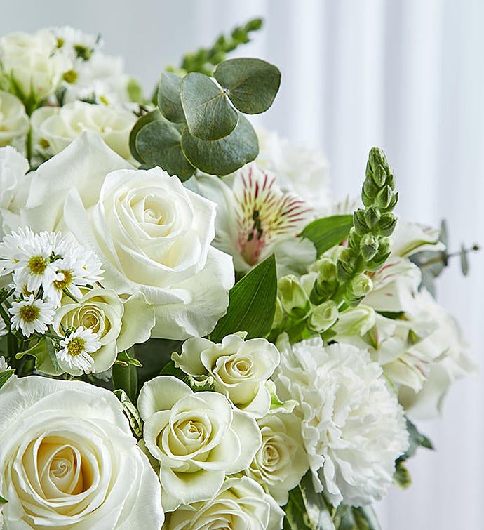 Zoomed in picture of Elegant all-white sympathy floral arrangement in a clear glass cylinder vase, featuring white roses, snapdragons, alstroemeria, carnations, and monte casino, accented with lush greenery and a Ti leaf ribbon. Perfect for memorials or as a thoughtful gift.