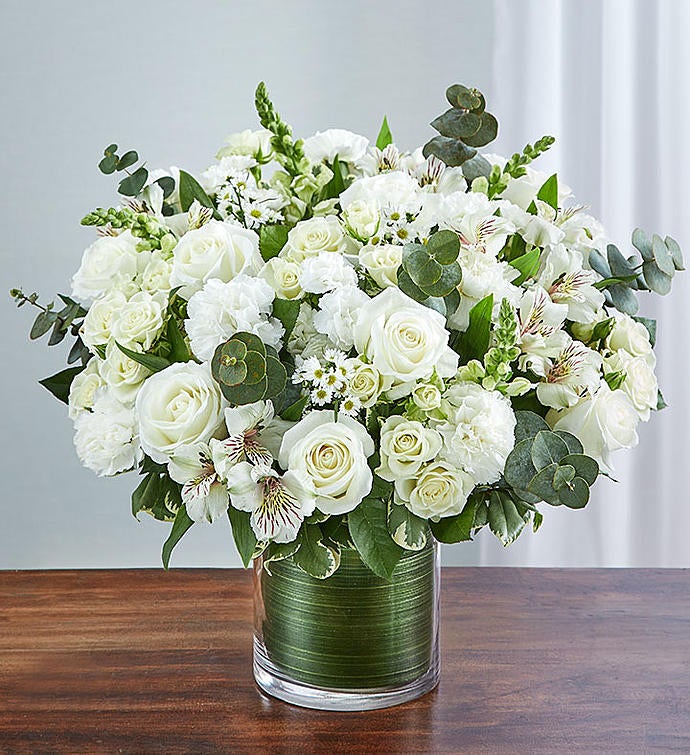 Large Elegant all-white sympathy floral arrangement in a clear glass cylinder vase, featuring white roses, snapdragons, alstroemeria, carnations, and monte casino, accented with lush greenery and a Ti leaf ribbon. Perfect for memorials or as a thoughtful gift.