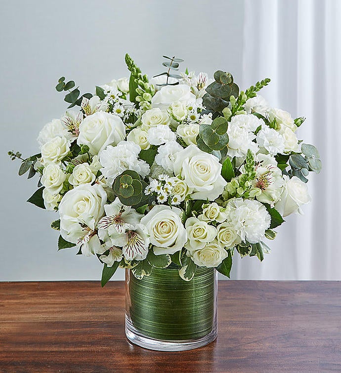 Medium Elegant all-white sympathy floral arrangement in a clear glass cylinder vase, featuring white roses, snapdragons, alstroemeria, carnations, and monte casino, accented with lush greenery and a Ti leaf ribbon. Perfect for memorials or as a thoughtful gift.