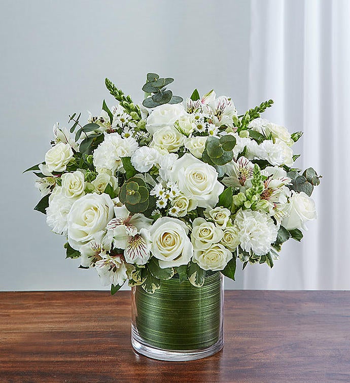 Small Elegant all-white sympathy floral arrangement in a clear glass cylinder vase, featuring white roses, snapdragons, alstroemeria, carnations, and monte casino, accented with lush greenery and a Ti leaf ribbon. Perfect for memorials or as a thoughtful gift.