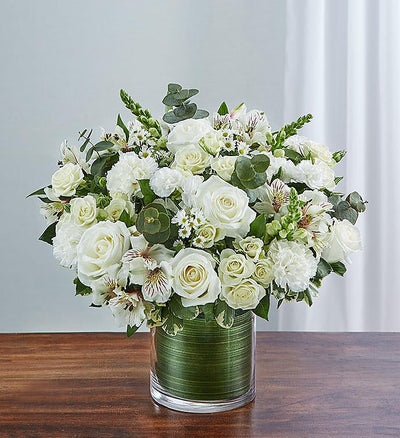 Small Elegant all-white sympathy floral arrangement in a clear glass cylinder vase, featuring white roses, snapdragons, alstroemeria, carnations, and monte casino, accented with lush greenery and a Ti leaf ribbon. Perfect for memorials or as a thoughtful gift. Small Elegant all-white sympathy floral arrangement in a clear glass cylinder vase, featuring white roses, snapdragons, alstroemeria, carnations, and monte casino, accented with lush greenery and a Ti leaf ribbon. Perfect for memorials or as a thoughtful gift.