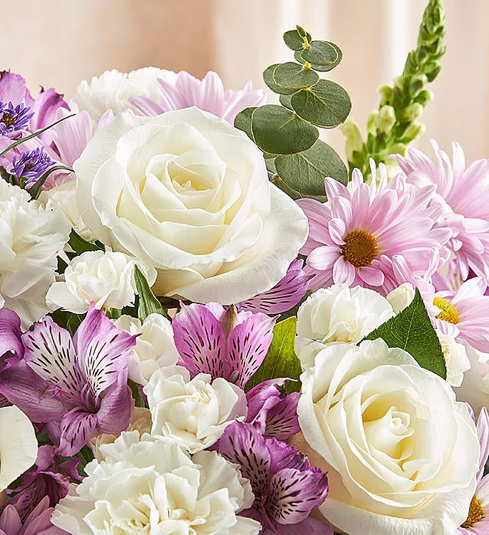 Zoomed in picture of Sympathy floral arrangement in lavender and white, featuring lavender roses, alstroemeria, carnations, white snapdragons, and daisy poms in a clear glass cylinder vase lined with a Ti leaf ribbon. A touching tribute for remembrance, perfect for home or service.