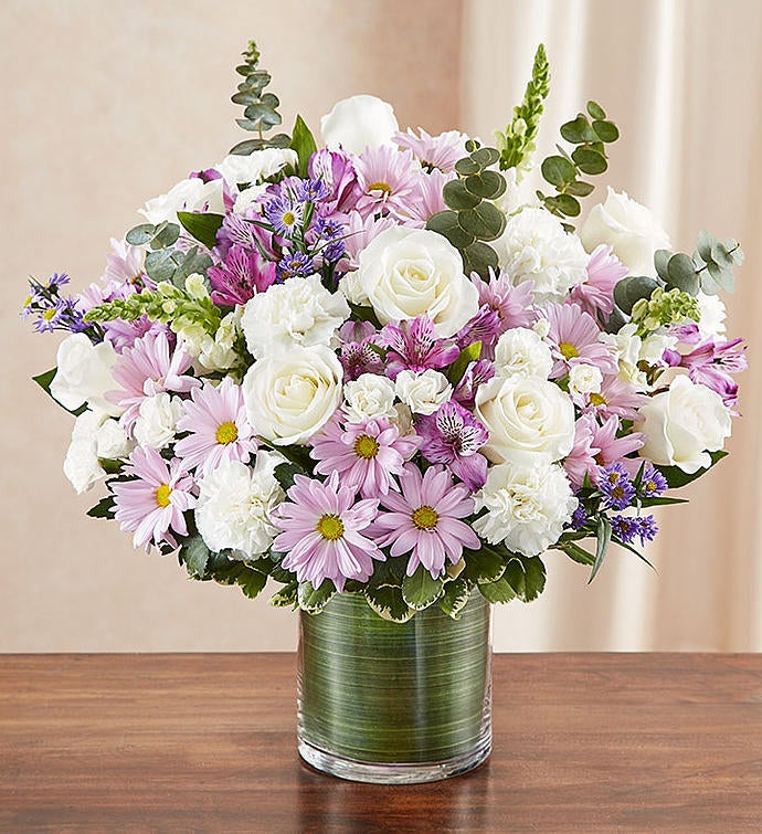 Large Sympathy floral arrangement in lavender and white, featuring lavender roses, alstroemeria, carnations, white snapdragons, and daisy poms in a clear glass cylinder vase lined with a Ti leaf ribbon. A touching tribute for remembrance, perfect for home or service.