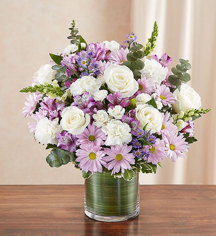 Medium Sympathy floral arrangement in lavender and white, featuring lavender roses, alstroemeria, carnations, white snapdragons, and daisy poms in a clear glass cylinder vase lined with a Ti leaf ribbon. A touching tribute for remembrance, perfect for home or service.