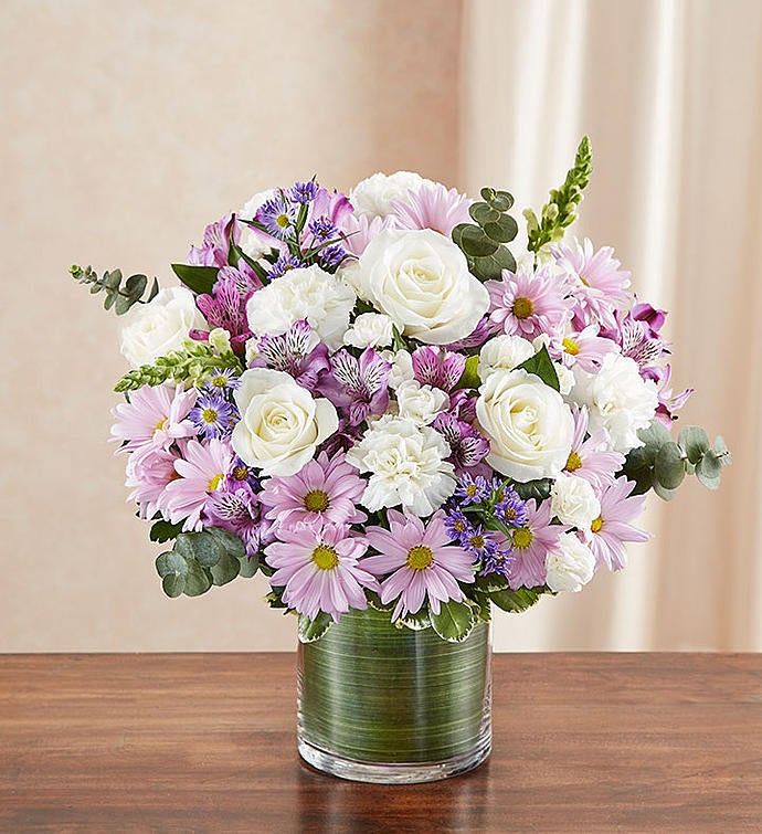 Small Sympathy floral arrangement in lavender and white, featuring lavender roses, alstroemeria, carnations, white snapdragons, and daisy poms in a clear glass cylinder vase lined with a Ti leaf ribbon. A touching tribute for remembrance, perfect for home or service.