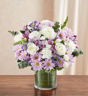 Small Sympathy floral arrangement in lavender and white, featuring lavender roses, alstroemeria, carnations, white snapdragons, and daisy poms in a clear glass cylinder vase lined with a Ti leaf ribbon. A touching tribute for remembrance, perfect for home or service.