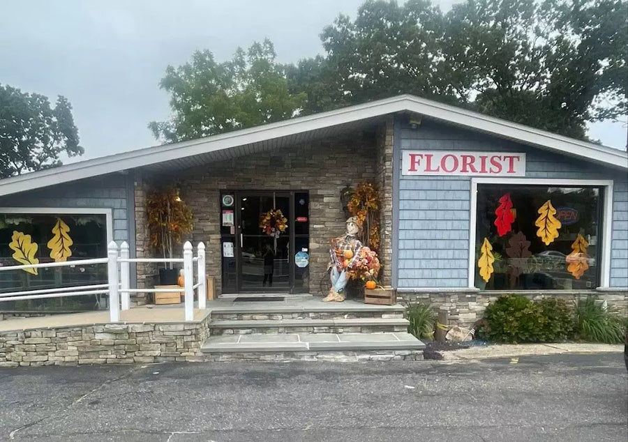 Front exterior of James Cress Florist Port Jefferson location, featuring a stone and blue siding storefront with seasonal fall decorations and large display windows showing floral arrangements.
