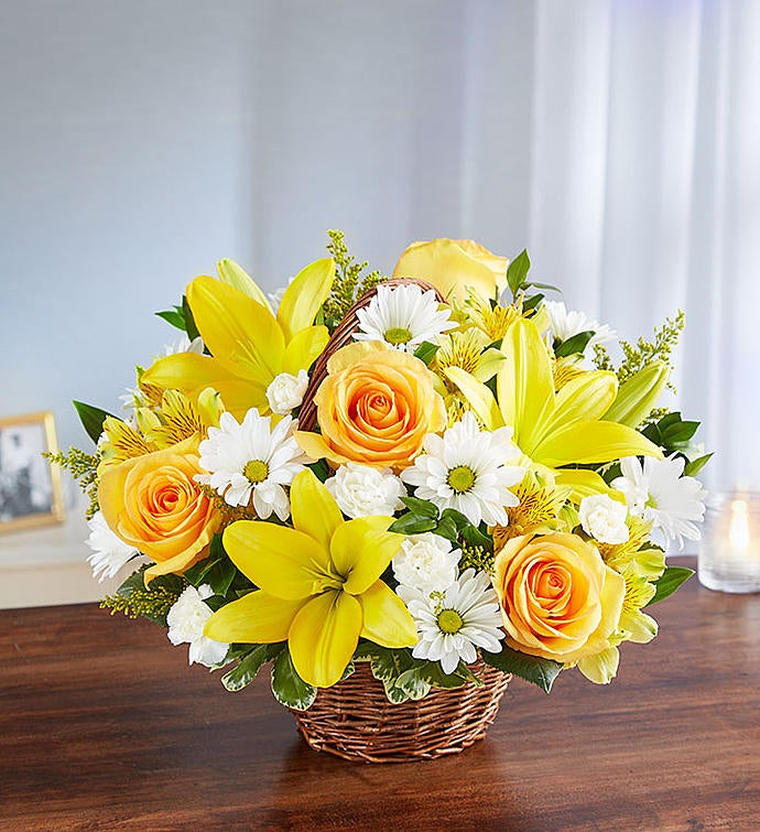 Medium yellow and white floral arrangement in wicker basket featuring yellow roses, Asiatic lilies, alstroemeria, white daisy poms, and mini carnations with lush greenery, perfect for conveying support and blessings.