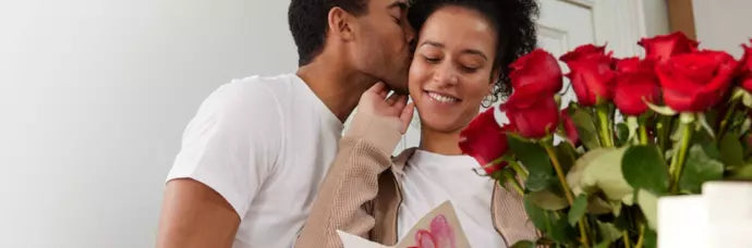 A smiling woman holding a card as a man kisses her cheek, with a bouquet of vibrant red roses in the foreground